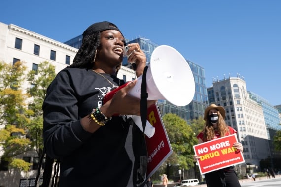 Person uses megaphone at a protest. Behind them, someone holds a sign that reads, "No More Drug War."