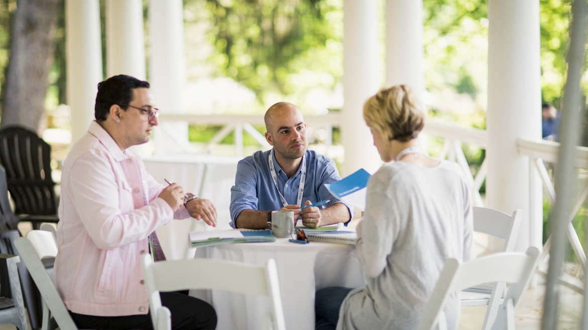 Three people wearing conference lanyards in conversation at a small table