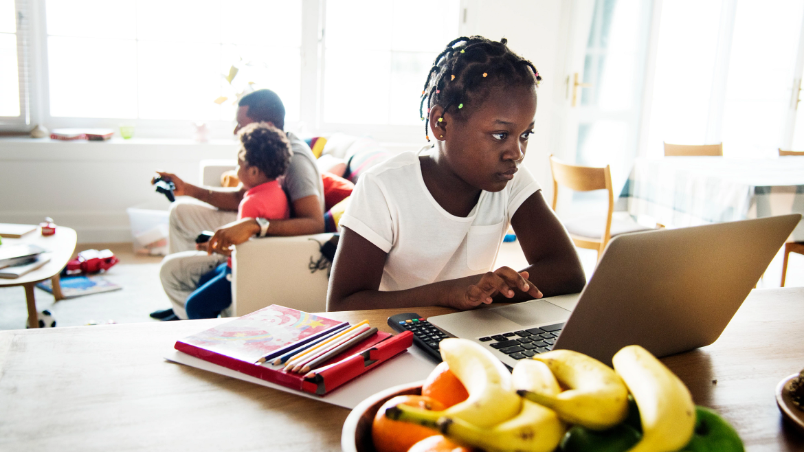 Girl studies at her laptop while father and younger child play video games behind her.