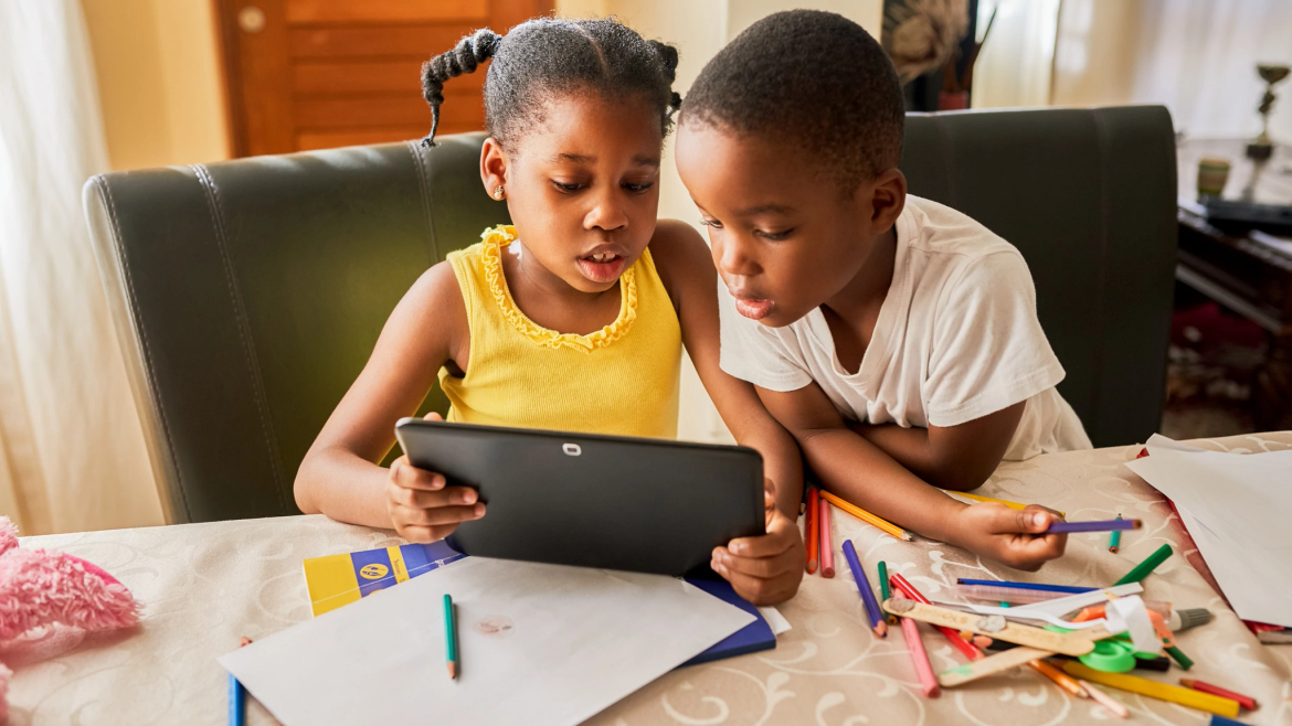 Two young children sharing a tablet screen. School supplies on the table.