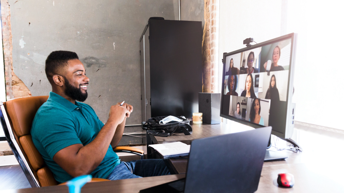 Man in a modern office smiling on a video call with others seen on desktop screen.