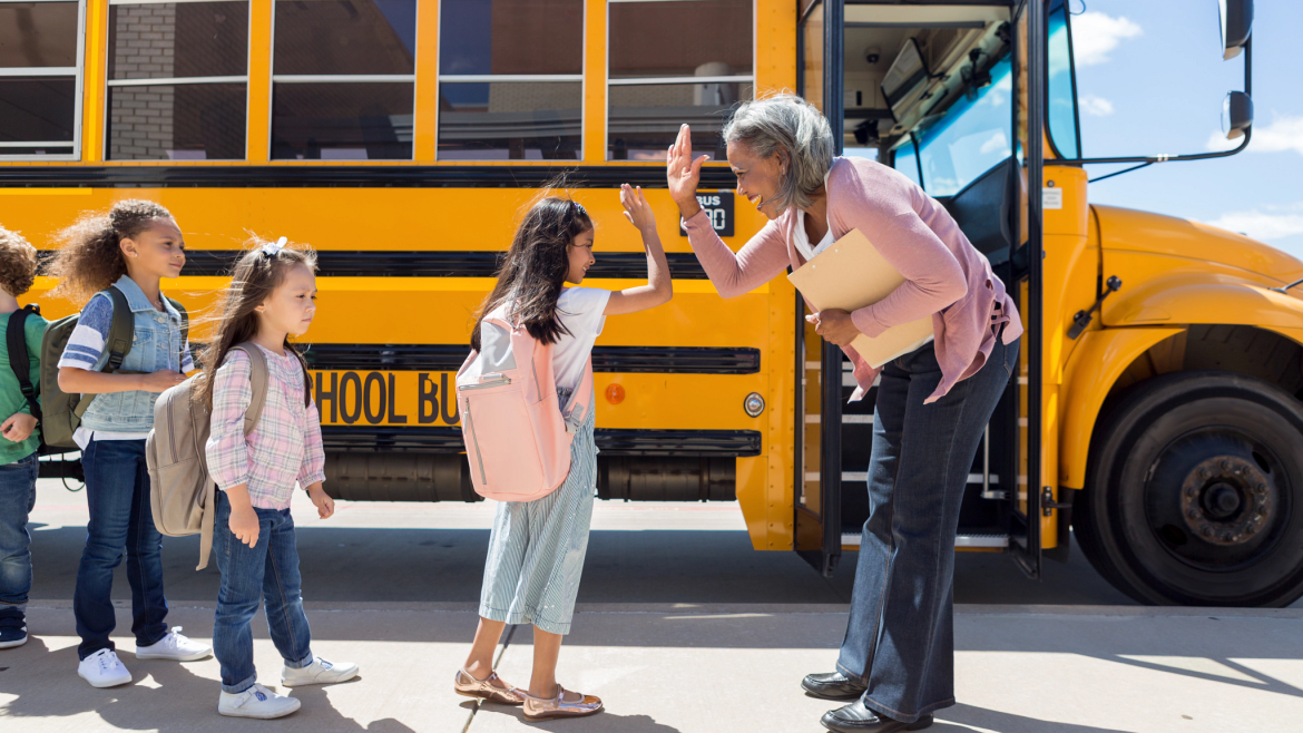 Teacher high-fives a little girl getting on a school bus as other kids wait their turn to board