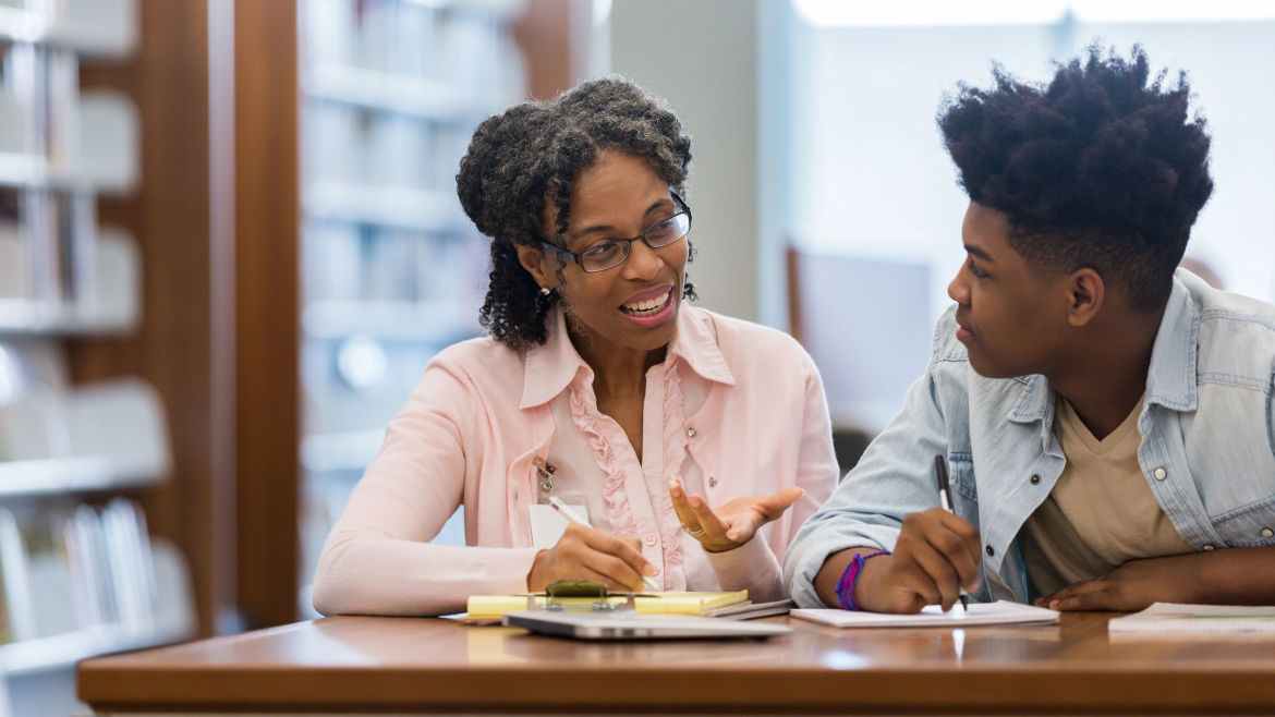 Teacher and high school student seated at library table She is smiling as she explains something to him.