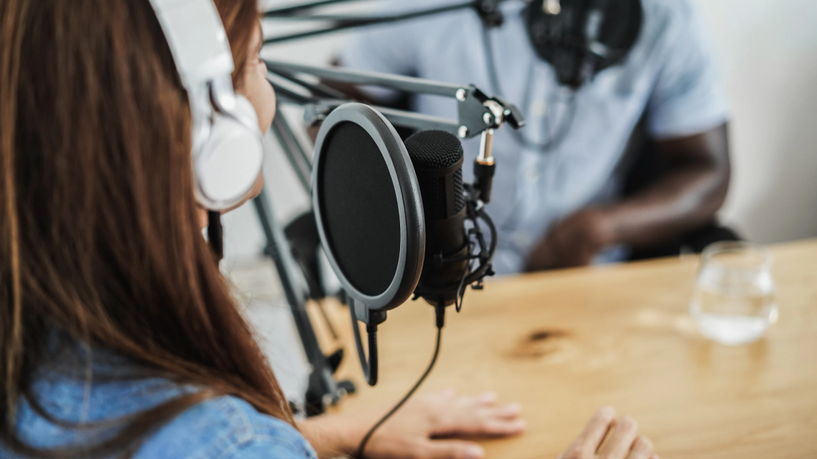 Two people wearing headphones sit at a table speaking into microphones