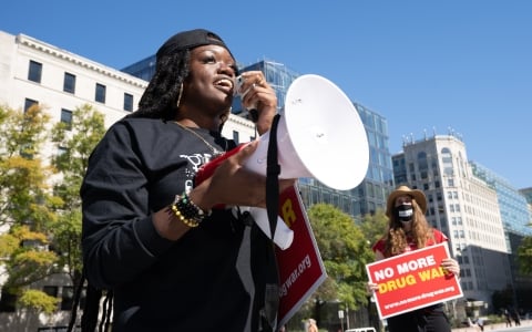 Person uses megaphone at a protest. Behind them, someone holds a sign that reads, "No More Drug War."
