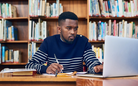Man with pen taking notes while reading off laptop screen